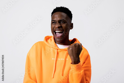 Hapy delighted black african american young guy in orange pullover hoodie whosing different emotions shaking fists closing eyes celebrating success celebrating victory different emotions. photo