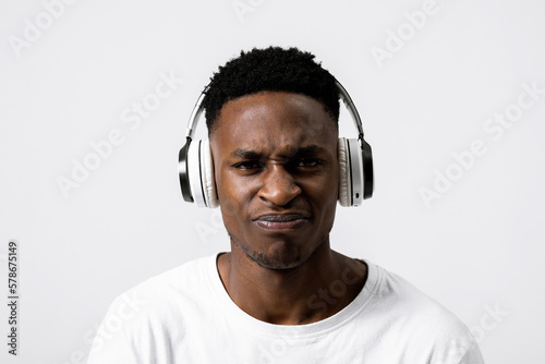 Sad dissatisfied african american black guy in white headphones listening to music news standing over grey background posing experssing different bad emotions.