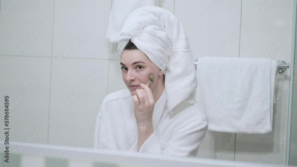 A young woman in a white towel massages herself with a jade roller. A young woman in a white towel massages herself with a jade roller.