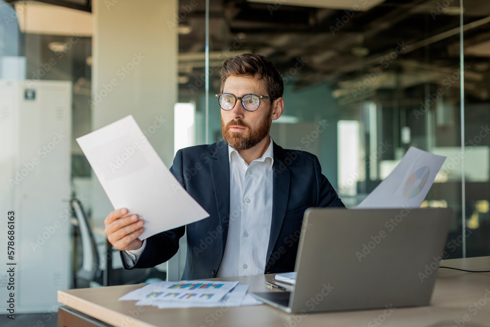 Shocked mature businessman reading documents, having problem with reports, working in office with laptop