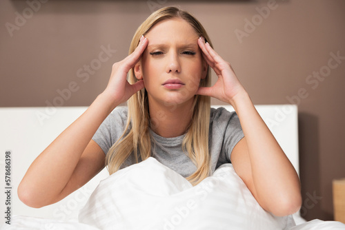 Pretty young blond woman lady in bedroom sitting on bed leaning on hand suffering from migraine pain having headache illness. Stressgul girl after hard day.