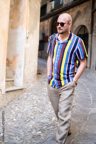 A man in a colorful shirt and loose pants enjoys his vacation in the south of Italy. He strolls through the streets and has an old black camera with him. He looks relaxed and in a good mood.