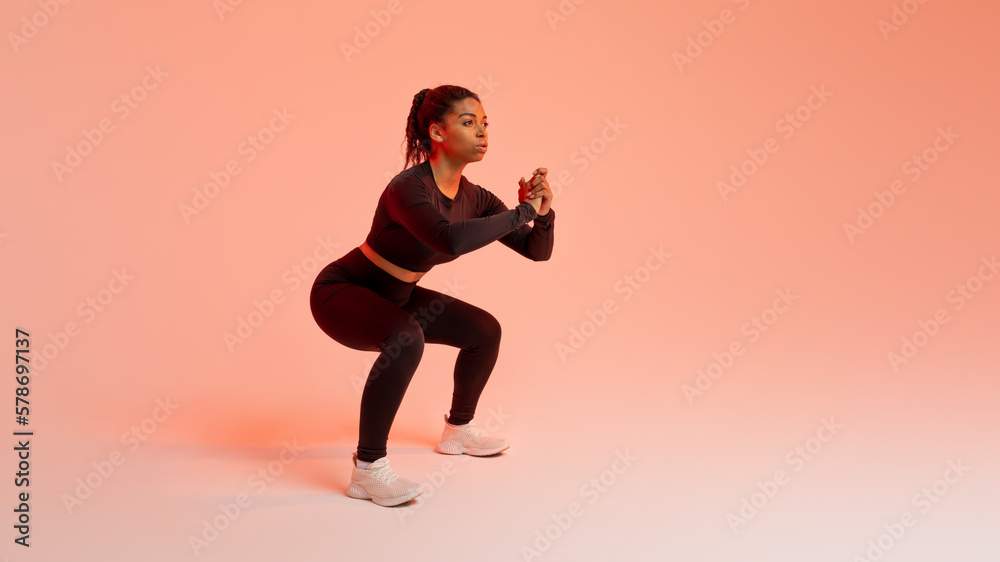 Training concept. Fit african american woman doing squats exercises, working out over peach neon background, free space