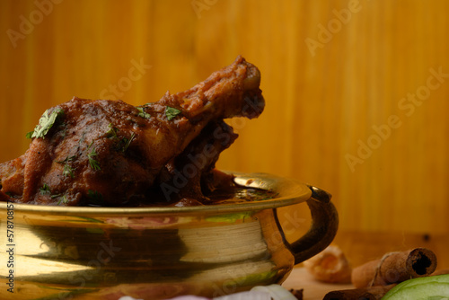 Spicy Reddish saoji Chicken Curry  Masala served in brass bowl or Kadhai ,with prominent Leg Piece, different spices and salad like cucumber onion carrot over  wooden background. photo
