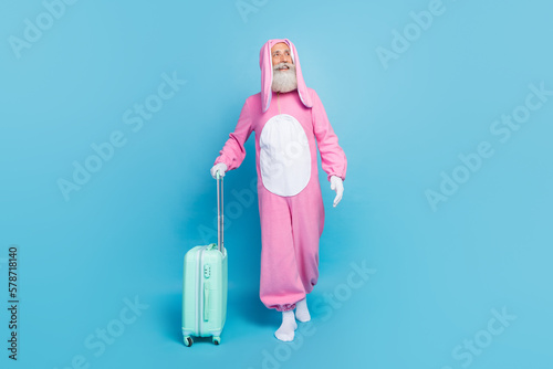 Full size portrait of cheerful aged man walking look empty space carry suitcase isolated on blue color background photo