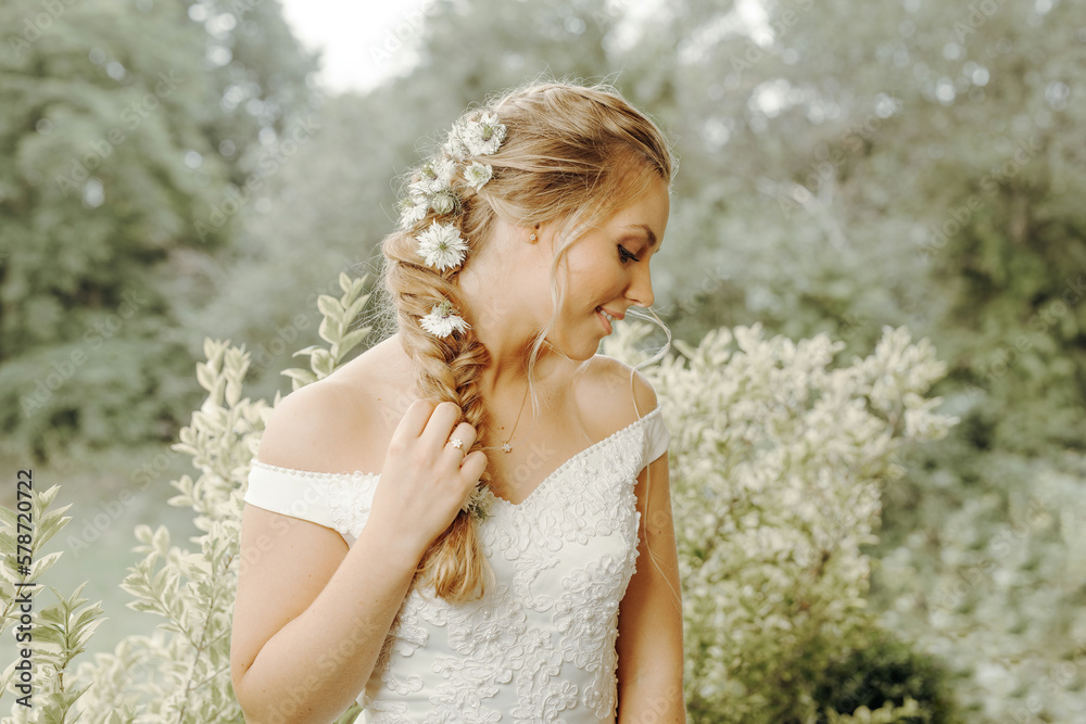 Beautiful woman with blond hair tied in a braid, decorated with flowers, is smiling and looking down, wearing a white dress, plants and trees in the background Beautiful woman with blond hair tied in a braid, decorated with flowers, is smiling and looking down, wearing a white dress, plants and trees in the background