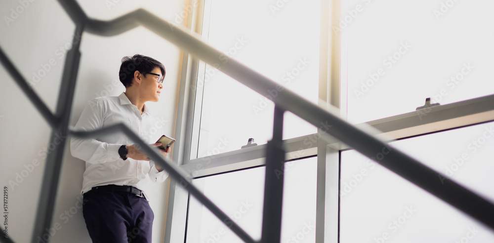 Handsome office worker hold mobile phone in hand looking out through the balcony see through glass window feeling lonely, miss girlfriend at office fire escape in relax time. Handsome office worker hold mobile phone in hand looking out through the balcony see through glass window feeling lonely, miss girlfriend at office fire escape in relax time.