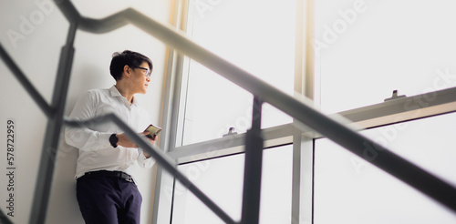 Handsome office worker hold mobile phone in hand looking out through the balcony see through glass window feeling lonely, miss girlfriend at office fire escape in relax time.