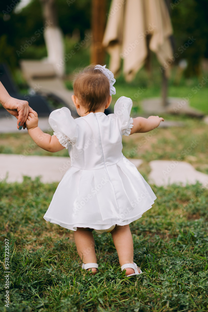 Adorable baby girl in white dress walking outdoors on grass with her mother holding her hand