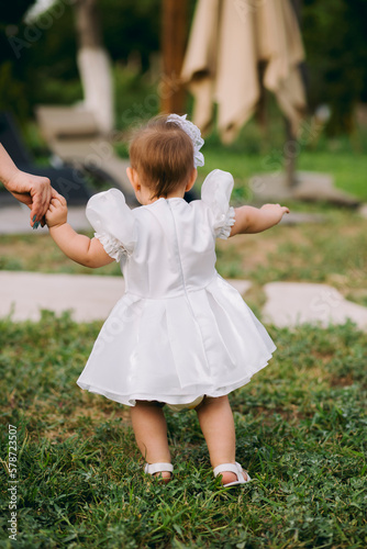 Obraz na płótnie Adorable baby girl in white dress walking outdoors on grass with her mother holding her hand w salonie
