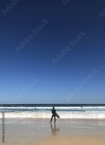 A surfer carrying her surfboard, standing on the beach with big beautiful blue sky, wave and sea in the background