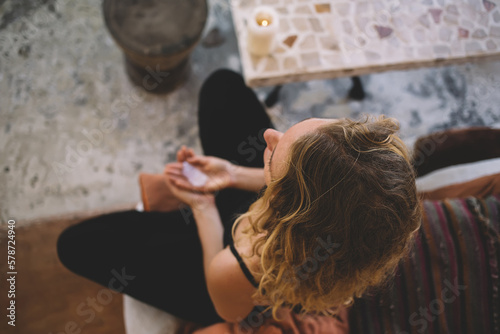 Young anonymous woman sitting on sofa with crystal and meditating