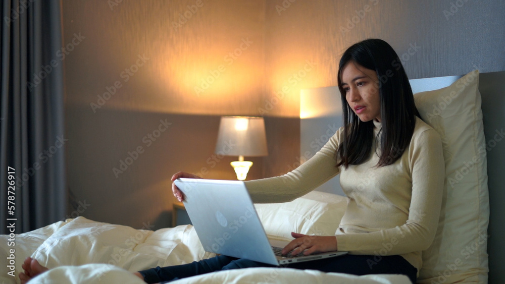 Young woman sitting on bed using laptop for work in bedroom at home.
