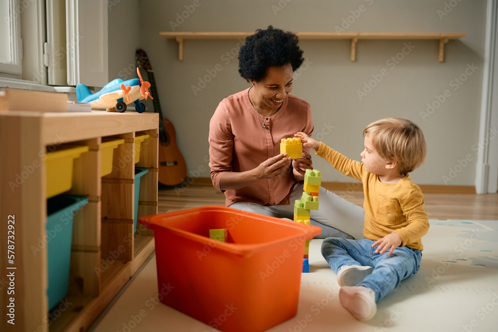 Happy black teacher and small boy play with toy blocks at kindergarten,