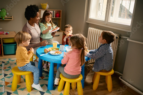 Group of children having lunch with their black female teacher at kindergarten.