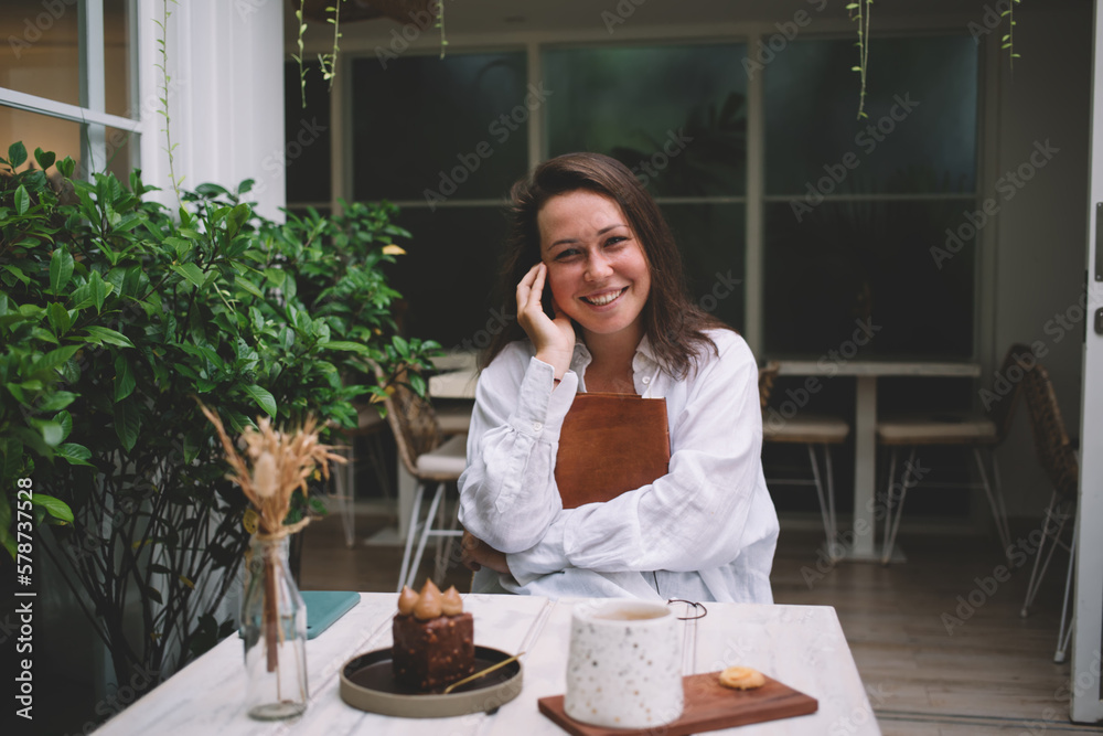 Half length portrait of cheerful female student with education textbook for learning and studying smiling at camera, happy Caucasian hipster girl spending weekend leisure in street coffee shop