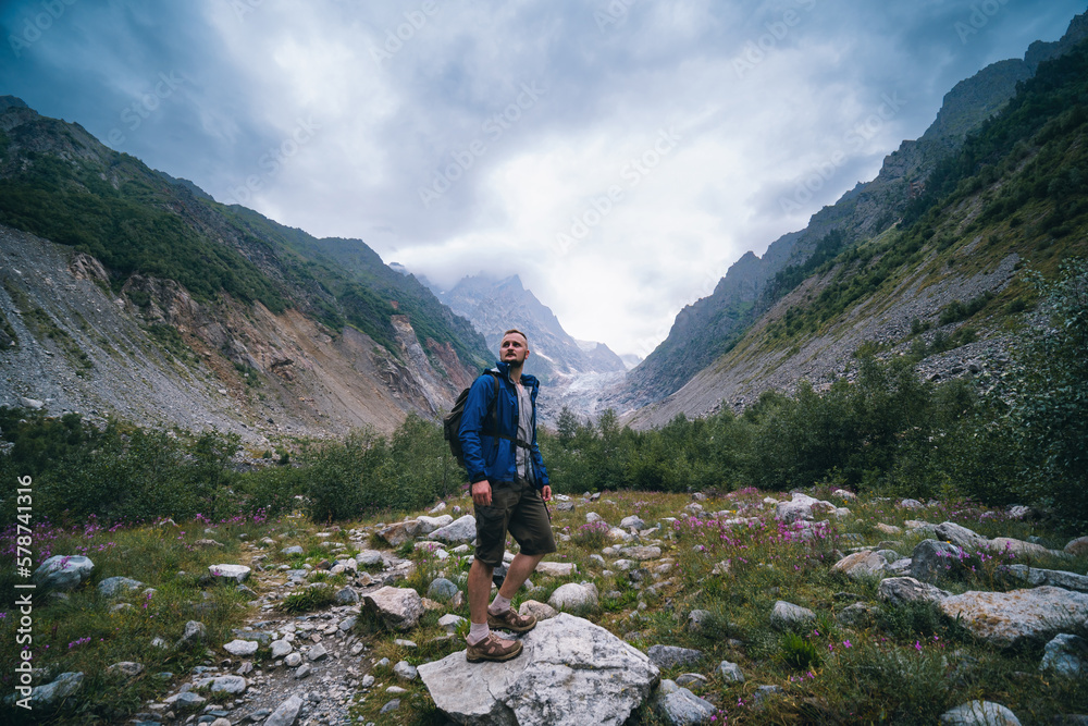 Traveler man with a backpack standing on a stone, On the way, walk the track. A valley among majestic high mountains.. Back view. The Chalaadi Glacier, Georgia, Caucasus. Blue jacket.