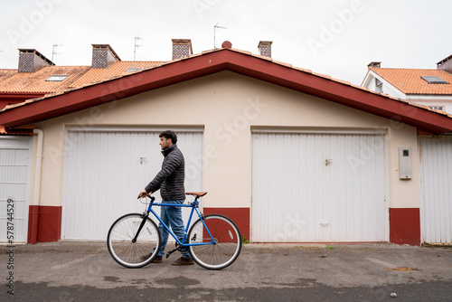 Man wearing jeans and black coat fixing a blue bike in front of garage