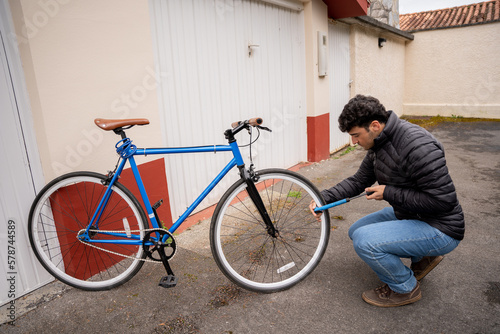 Man wearing jeans and black coat fixing a blue bike in front of garage