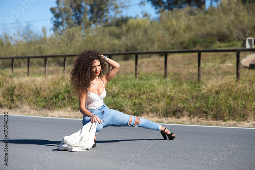 beautiful brunette woman with curly hair is standing in the middle of the road with her leg stretched out. The woman is dressed in jeans torn at the knee and white lingerie.