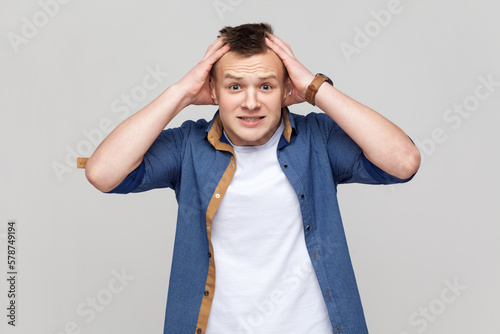 Teenager boy wearing blue shirt keeping hands on head and looking frighten with frightened eyes.
