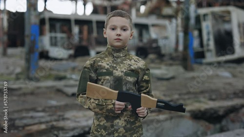 Portrait of the Serious Little Soldier Kid Holding Toy Gun in the Hands and Standing in the Middle of Destroyed Plant. Ghetto. Boy Warrior Playing War photo