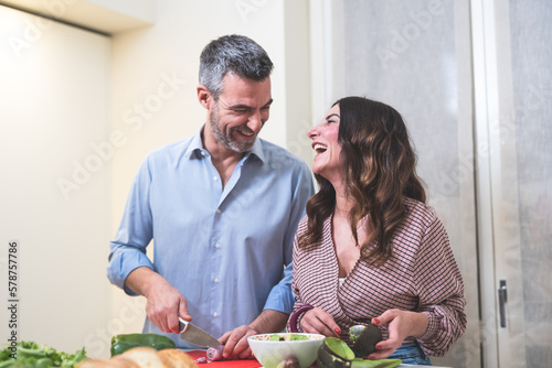 Happy Couple Preparing Healthy Food and similing together .Adult man with hair grey chops spring onion while woman prepares avocado to cook guacamole photo