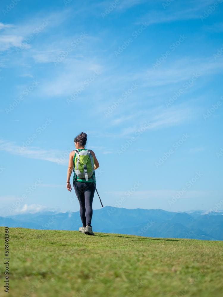 mujer de espaldas caminando por la montaña encima de la hierba con cielo azul llevando una mochila