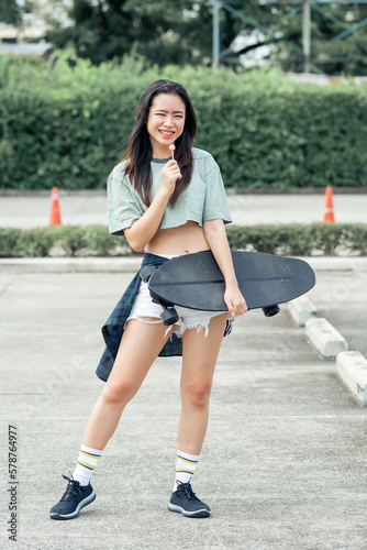 Portrait beautiful sportive Asian female skater wearing hipster clothes with lollipop, smiling with happiness, holding skateboard and standing outdoor with copy space. Activity and Adventure Concept.