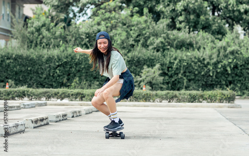 Portrait beautiful sportive Asian female skater wearing hipster shirt with shorts, smiling with happiness, standing on skateboard and playing outdoor with copy space. Activity and Adventure Concept.