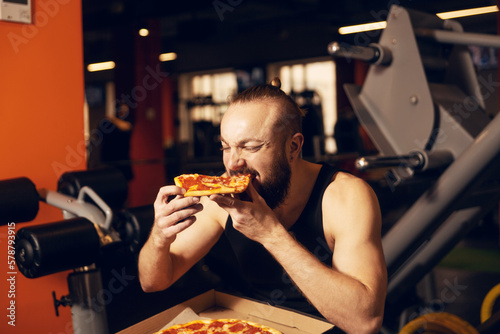 A young man with a beard is happy to eat pizza in the gym.