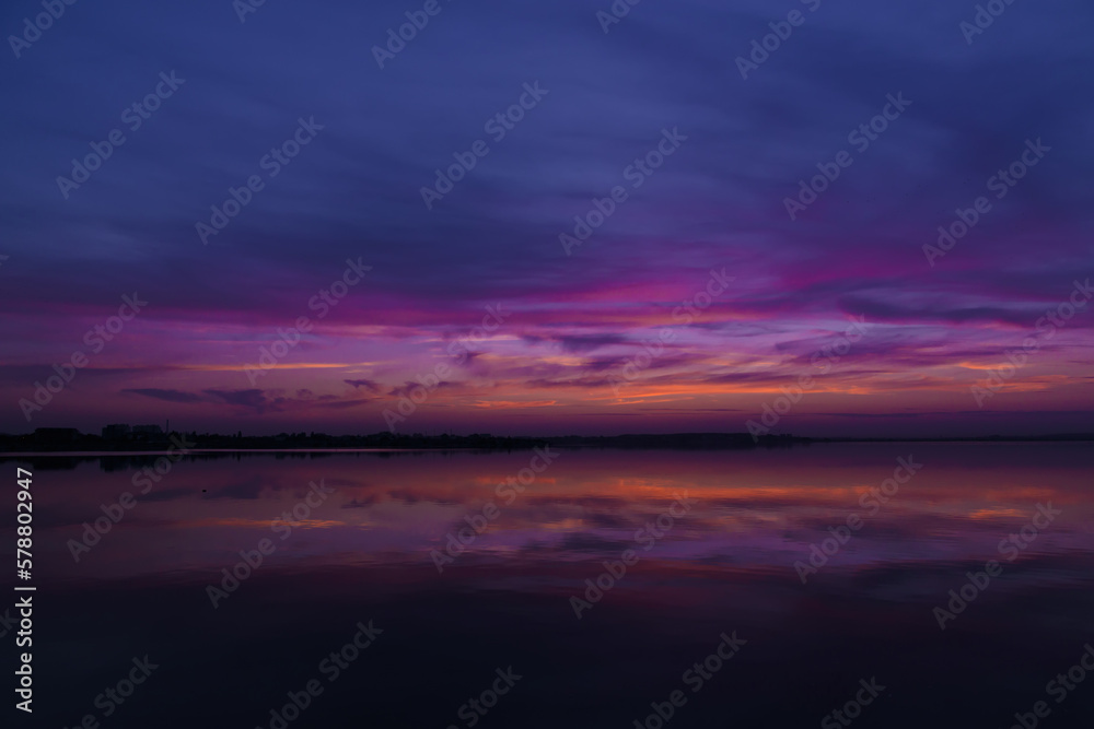 Scenic view of colorful sky reflected in the lake at evening, after sunset. Sunset at coast of the lake. Reflection, blue sky and yellow sunlight. Landscape during sunset. The beautiful nature around