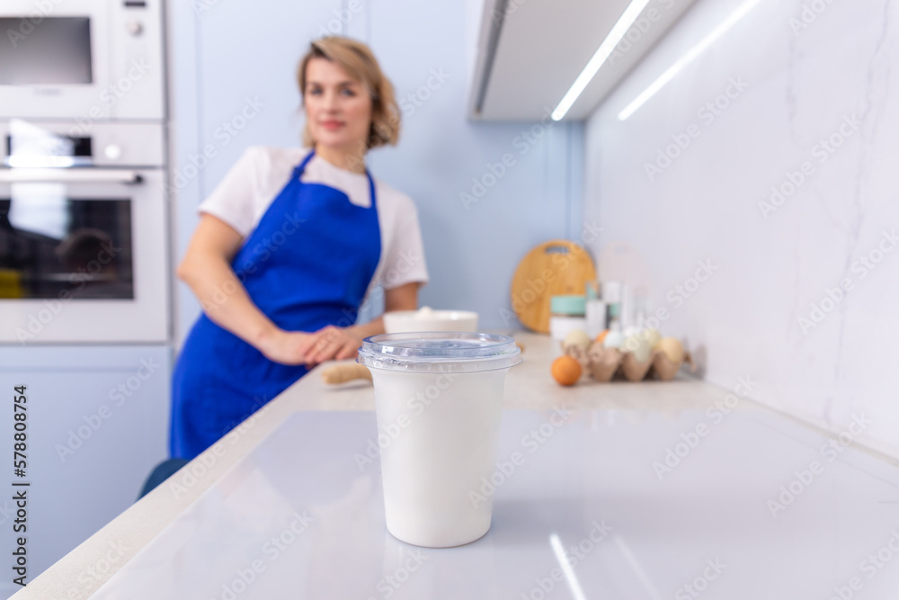 A beautiful young housewife poses in the kitchen against the background of a plastic cup with sour cream or yogurt. A beautiful young housewife poses in the kitchen against the background of a plastic cup with sour cream or yogurt.