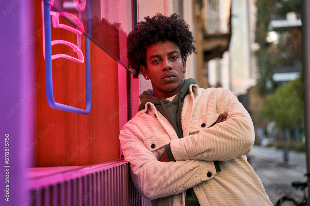Confident cool African American hipster guy standing at city street near neon light sign. Rebel gen z male model looking at camera outside advertising young generation culture life entertainment. - obrazy, fototapety, plakaty 
