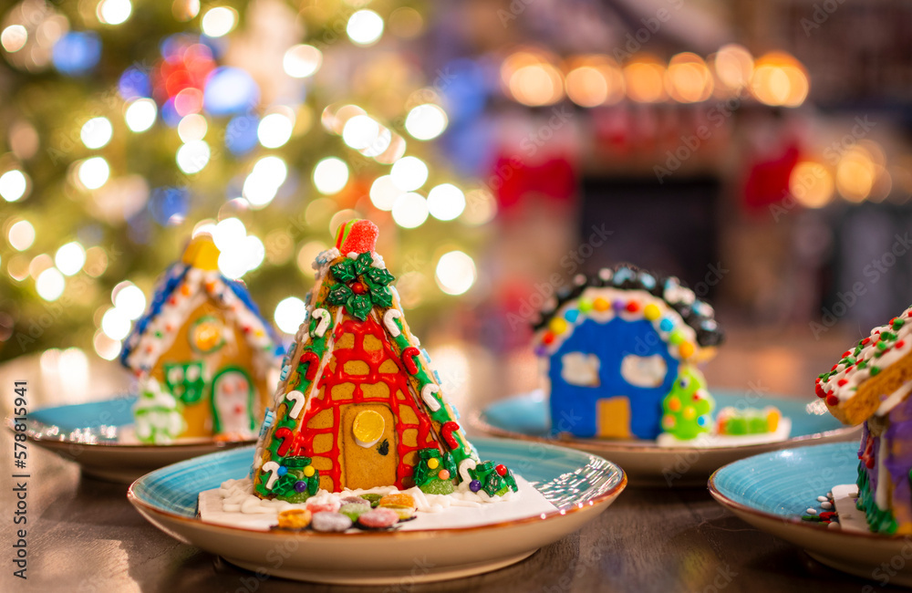 A colorful, frosted homemade gingerbread house with others behind with a lighted Christmas tree and fireplace with stockings on the mantel inside a home.