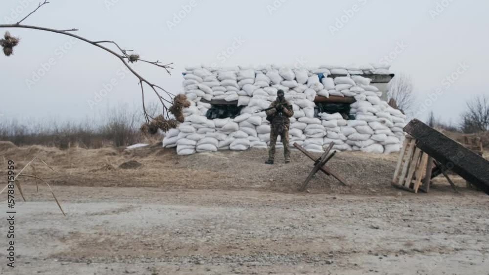 Soldier with weapon in military uniform stands in front of the barricades from sandbags and anti-tank hedgehog barriers. Military man on the roadblock. Combatant in full ammunition. Concept of war Soldier with weapon in military uniform stands in front of the barricades from sandbags and anti-tank hedgehog barriers. Military man on the roadblock. Combatant in full ammunition. Concept of war