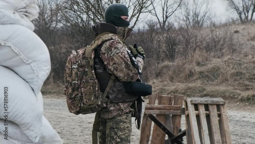 Soldier with weapon in military uniform stands in front of the barricades from sandbags and anti-tank hedgehog barriers. Military man on the roadblock. Combatant in full ammunition. Concept of war photo