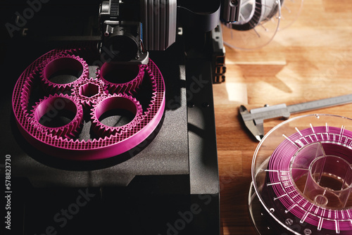 Black FDM-printer makes planetary gear machine part with visible infill from pink filament. Top view on printer, tools and filament roll on wooden desk area. Modern production concept. Selective focus