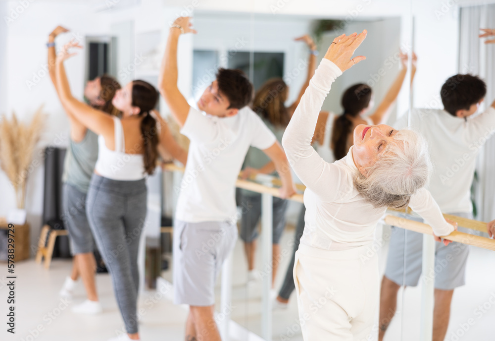 Fototapeta premium Gracile old-aged woman engaging in ballet at ballet barre in training hall during workout session
