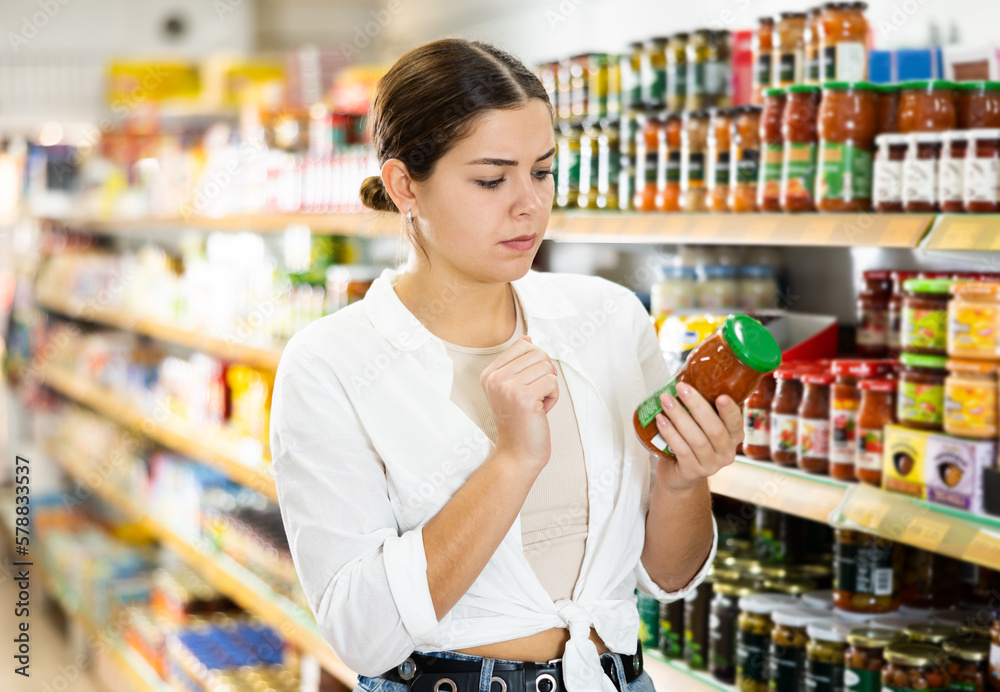 Interested thoughtful young female shopper choosing food in supermarket, holding glass jars with canned goods and reading labels..