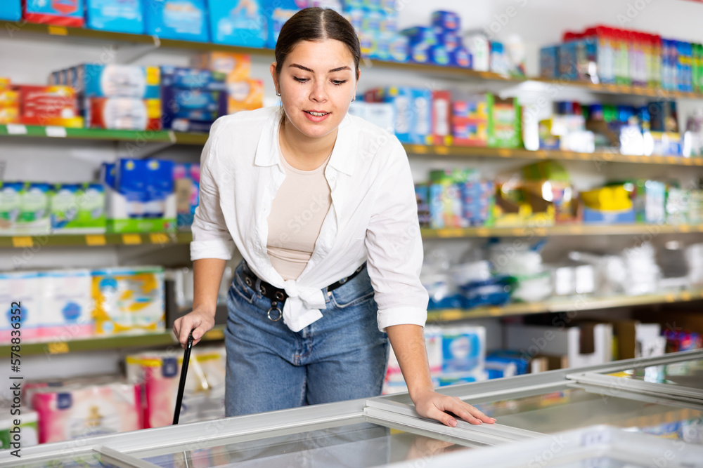 Interested emotional young woman standing near refrigerated display cases choosing frozen food in supermarket