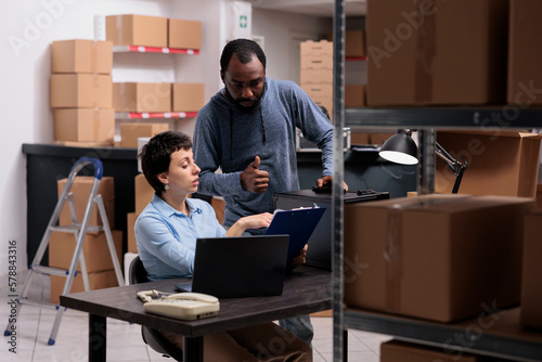 Supervisor woman analyzing metallic delivery order on clipboard discussing transportation logistics with worker while working together in warehouse. Storehouse team checking distribution stock