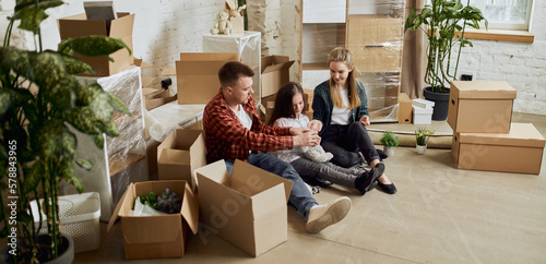 Young happy family, man, woman and kid sittin on floor in new living room. Moving into new flat, apartment with many cardboard boxes. Concept of moving houses, real estate, family, new life