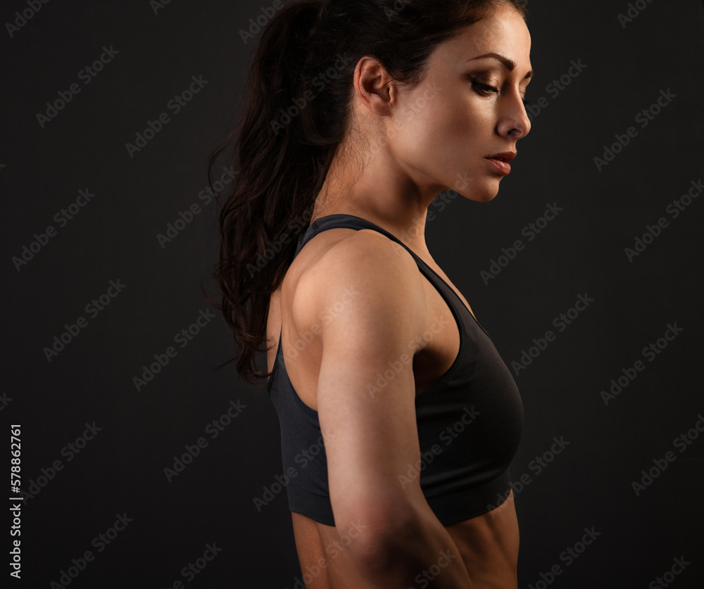 Serious female sporty muscular with ponytail doing stretching workout the shoulders, blades and arms in sport bra, standing on dark shadow grey background with empty copy space. Profile Serious female sporty muscular with ponytail doing stretching workout the shoulders, blades and arms in sport bra, standing on dark shadow grey background with empty copy space. Profile