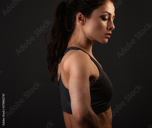 Serious female sporty muscular with ponytail doing stretching workout the shoulders, blades and arms in sport bra, standing on dark shadow grey background with empty copy space. Profile