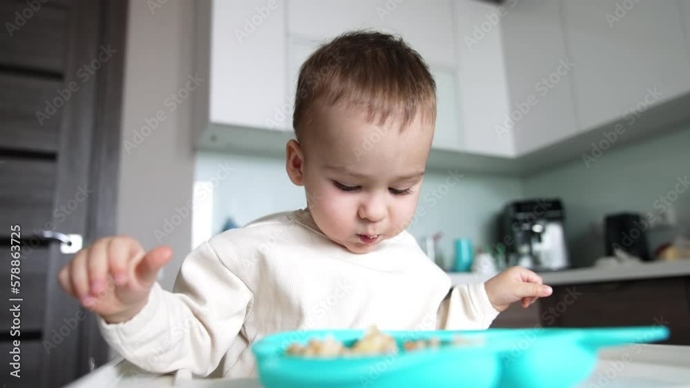 Beautiful Caucasian boy in white shirt having lunch. Mom’s hand takes some food with a spoon and gives it to her son. Low angle view. Beautiful Caucasian boy in white shirt having lunch. Mom’s hand takes some food with a spoon and gives it to her son. Low angle view.