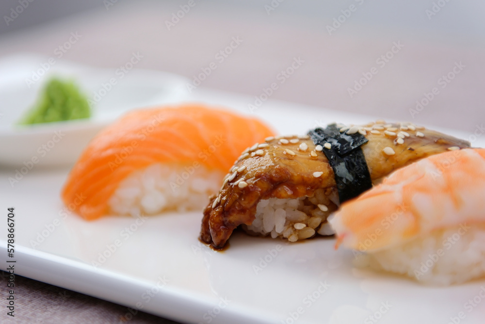 Closeup hand holding bamboo chopsticks with nigiri shrimp while soaking it in soy sauce. Detail of sushi set on wooden tray at restaurant while hand dip nigiri in soy sauce. Japanese cuisine concept Closeup hand holding bamboo chopsticks with nigiri shrimp while soaking it in soy sauce. Detail of sushi set on wooden tray at restaurant while hand dip nigiri in soy sauce. Japanese cuisine concept