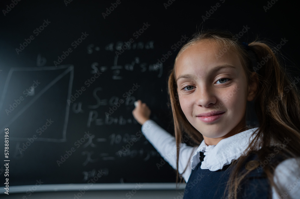 Portrait of a caucasian girl in the classroom. The schoolgirl writes the formula with chalk on the blackboard and looks into the camera. Portrait of a caucasian girl in the classroom. The schoolgirl writes the formula with chalk on the blackboard and looks into the camera.