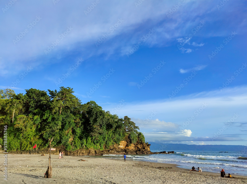 A beautiful beach with its sandy shore and waves, turquoise blue sea, green hills, and blue sky with clouds on a sunny day. It's a tropical summer view, a panoramic landscape