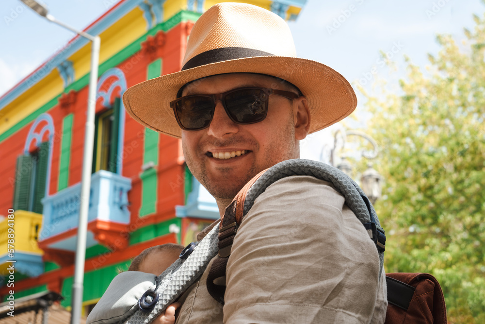 Caucasian happy man, father carrying little newborn baby in backpack,family traveling, going sightseeing in center of La Boca quarter, Buenos Aires, Argentine, looking at bright colourful buildings. Caucasian happy man, father carrying little newborn baby in backpack,family traveling, going sightseeing in center of La Boca quarter, Buenos Aires, Argentine, looking at bright colourful buildings.